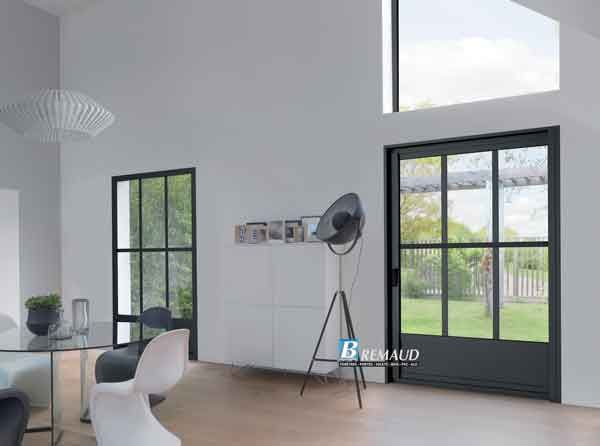 Vue intérieure : murs blancs, portes à cadre noir, table en verre, lampadaire, décoration moderne et vue sur la cour.