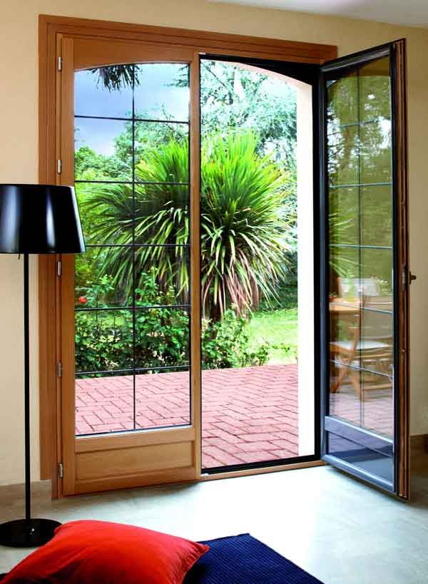 Des portes-fenêtres en bois s'ouvrent sur un jardin, avec une lampe noire et un coussin rouge au premier plan.