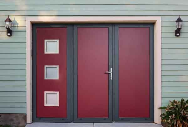 Porte de garage rouge et grise avec trois fenêtres dépolies, encadrée de moulures gris clair et de bardage vert.