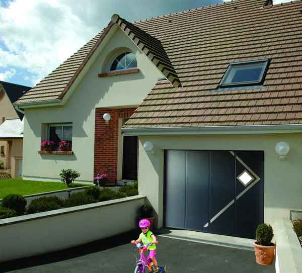 Un enfant fait du vélo rose dans une allée devant une maison dont la porte de garage est gris foncé.