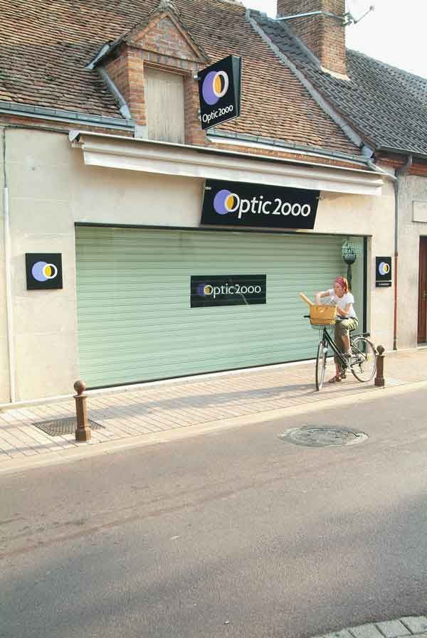 Magasin Optic 2000 aux volets verts fermés. Personne à vélo avec panier.