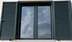 Fenêtre aux volets sombres ouverts, reflétant un ciel nuageux.