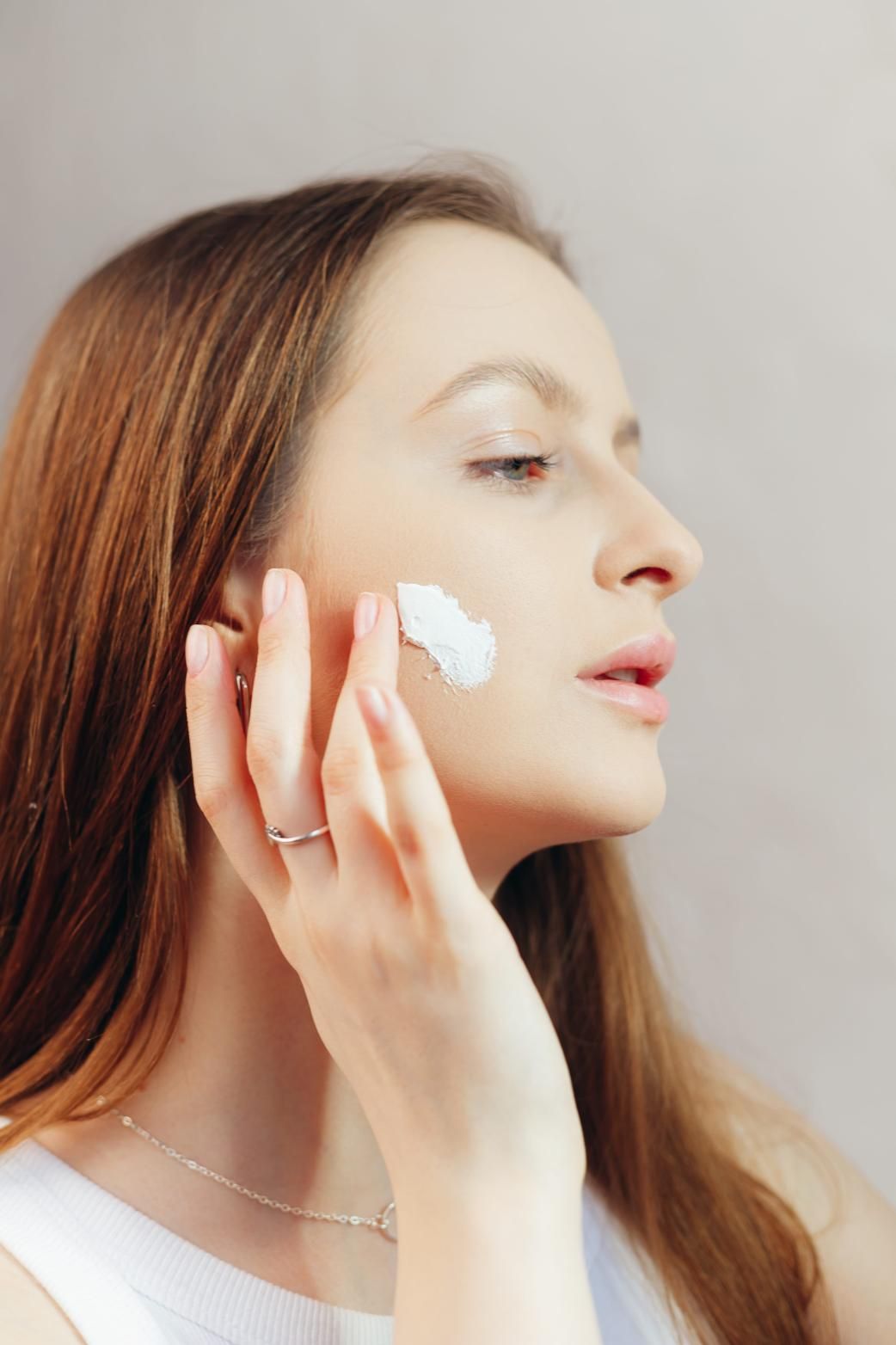 Mujer aplicándose crema blanca en la mejilla con un disco de algodón, perfil lateral sobre un fondo neutro.