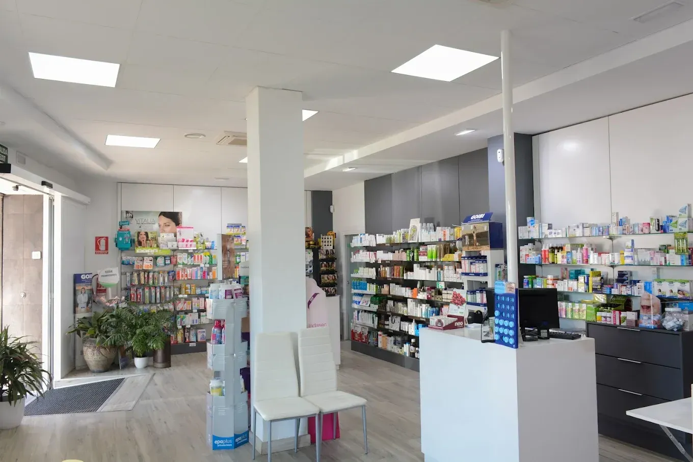 Interior luminoso de una farmacia con pasillos de medicamentos, estantes, mostrador y una columna de soporte blanca.