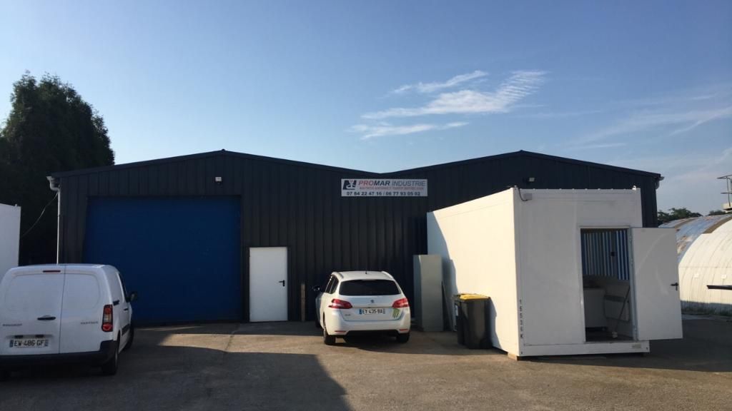 Un bâtiment commercial avec une porte bleue, une camionnette blanche, une voiture et un conteneur blanc ouvert, par une journée ensoleillée.