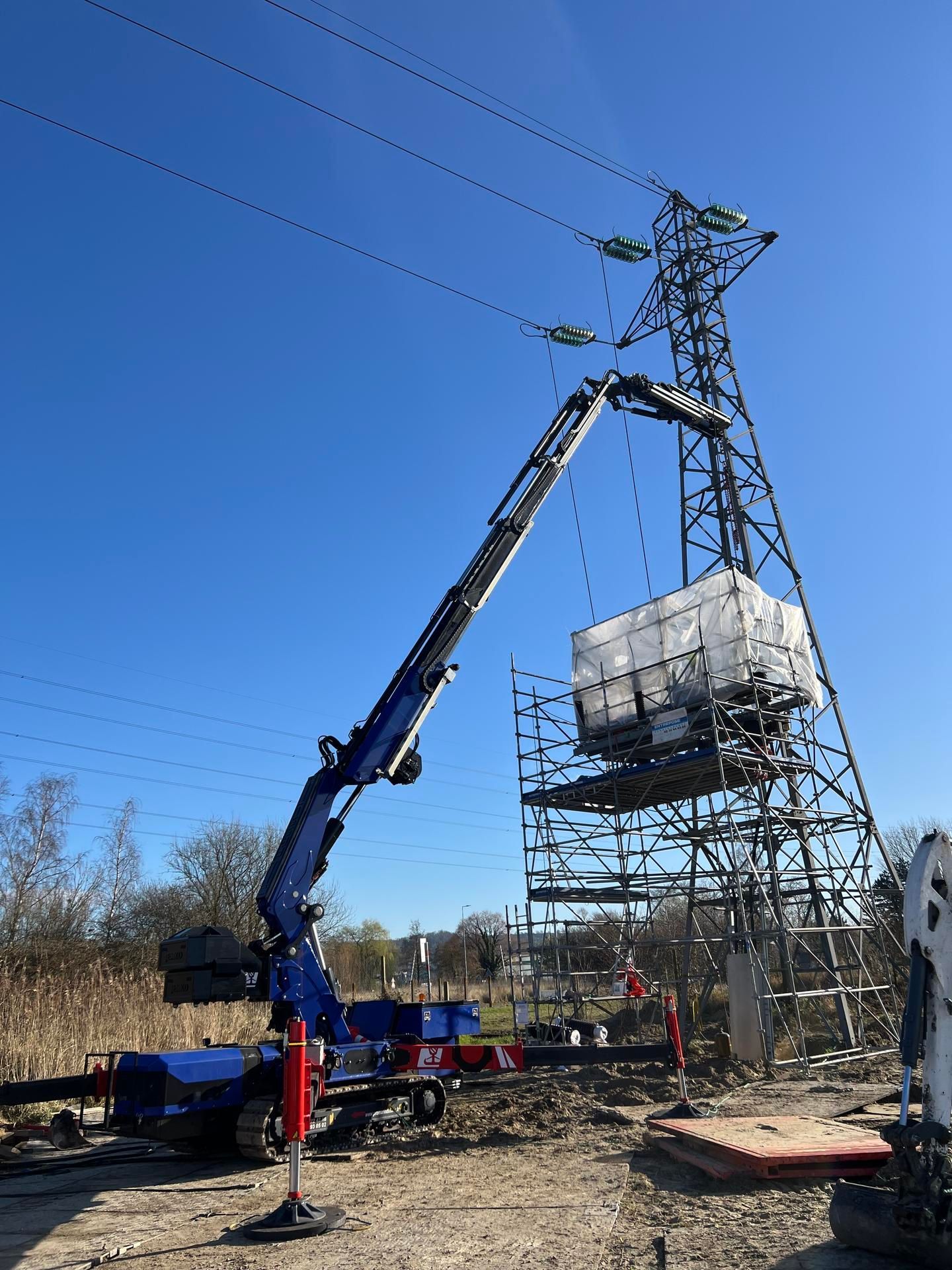 Une grue bleue soulève du matériel près d'un pylône à haute tension sous un ciel bleu.