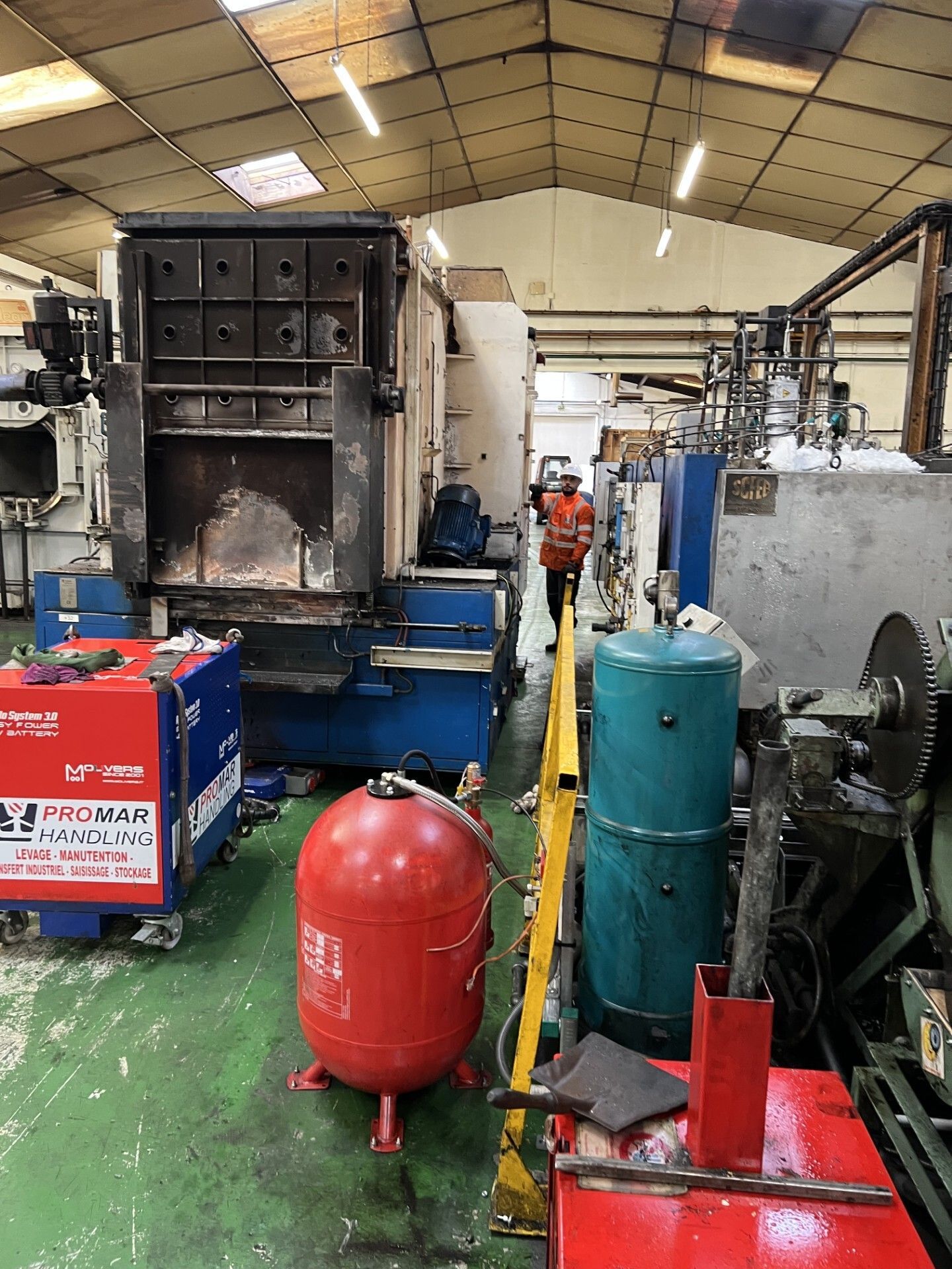 Atelier industriel avec de grosses machines, une cuve rouge et un ouvrier en gilet de sécurité orange.