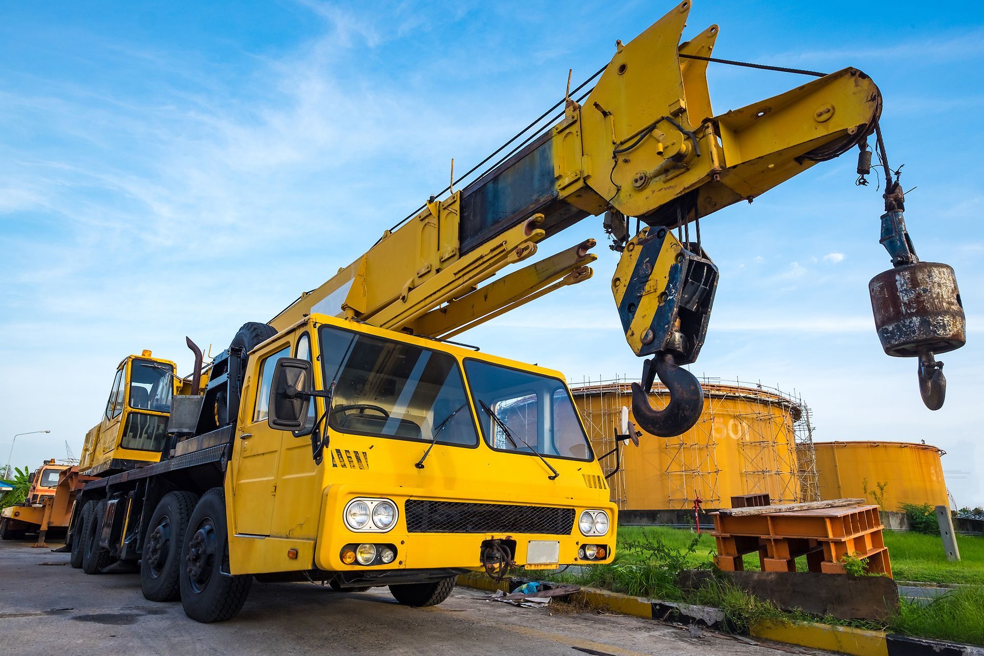 Grue mobile jaune sur une route près de réservoirs industriels, soulevant une charge lourde. Ciel bleu.