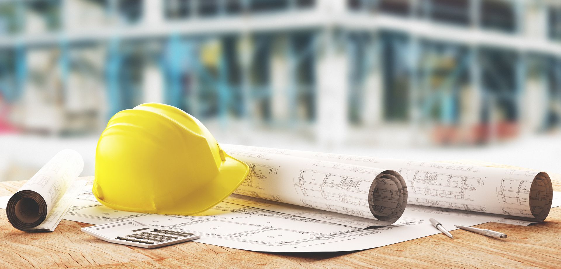 Un casque de chantier jaune et des plans roulés sur une table, avec un bâtiment en arrière-plan.