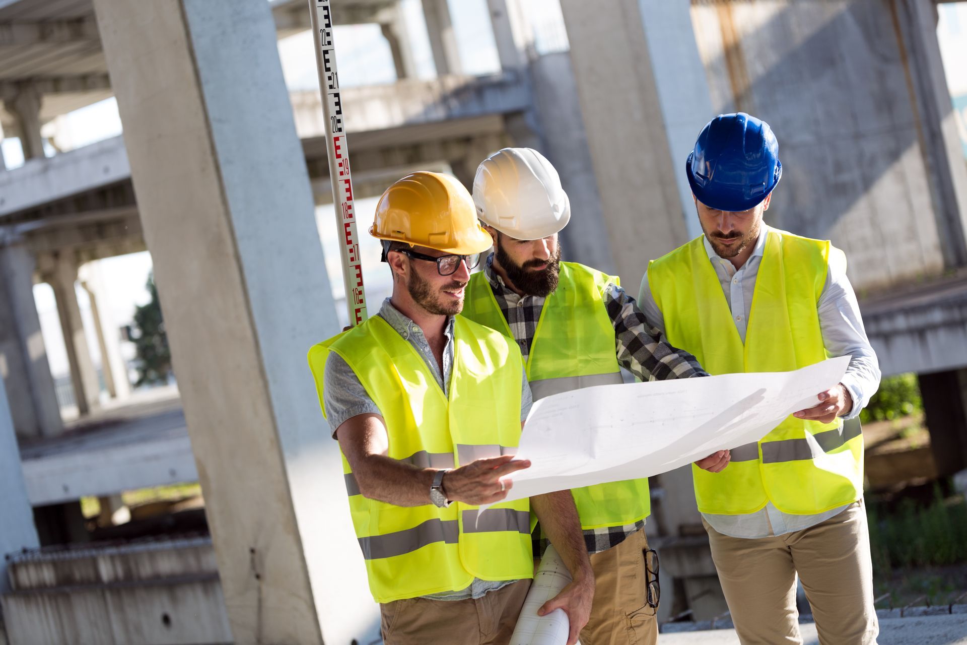 Trois ouvriers du bâtiment, casqués et vêtus de gilets de sécurité, examinent des plans sur un chantier.