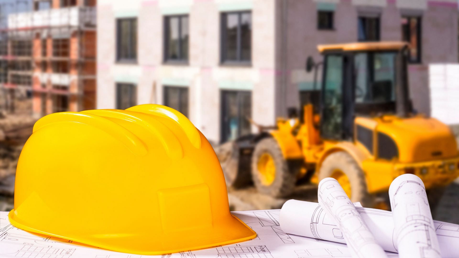 Casque jaune et plans sur un chantier ; engins de chantier et bâtiment flous.