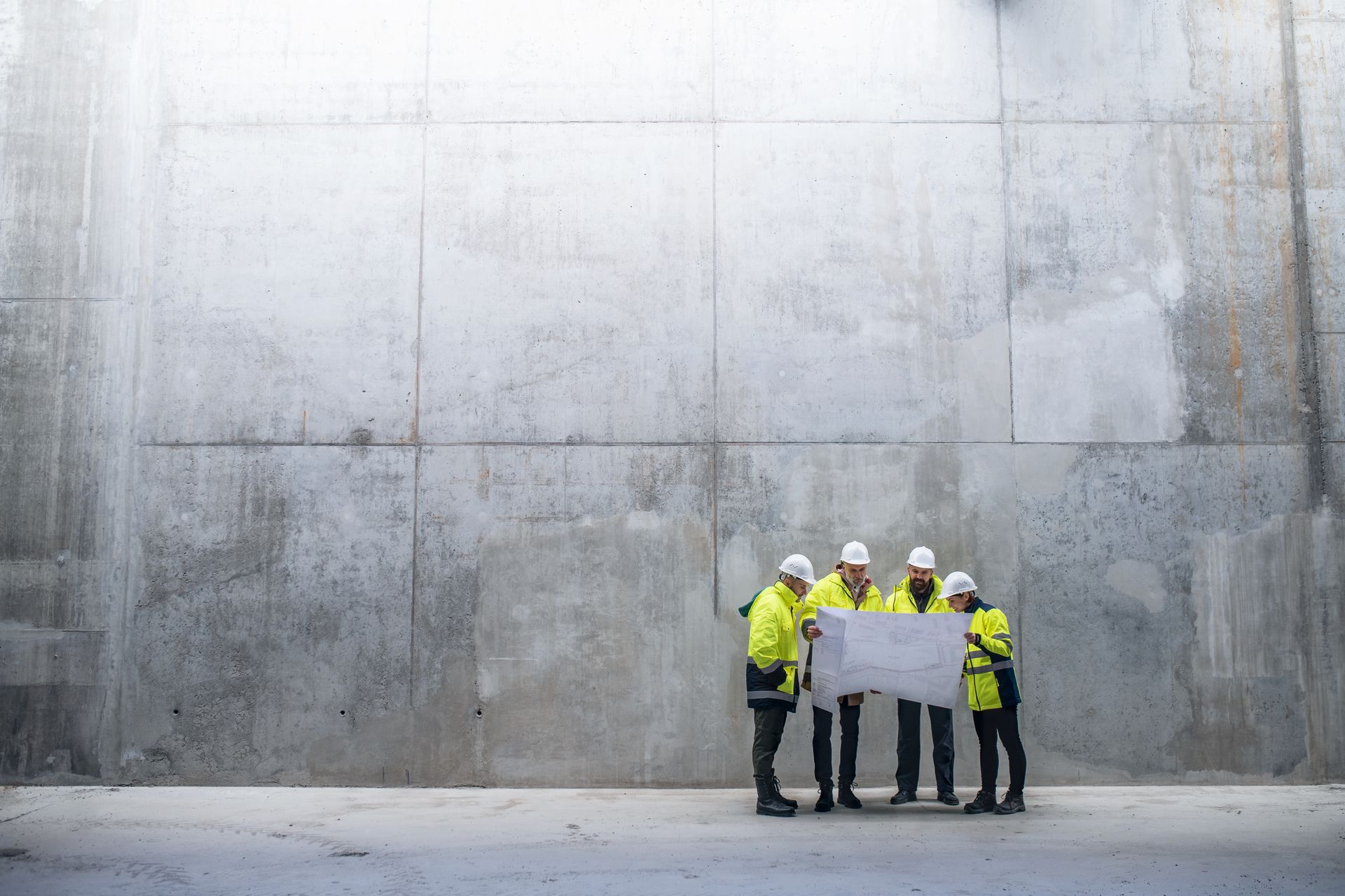 Quatre ouvriers du bâtiment, portant des gilets et des casques de sécurité, examinent un plan contre un grand mur de béton.
