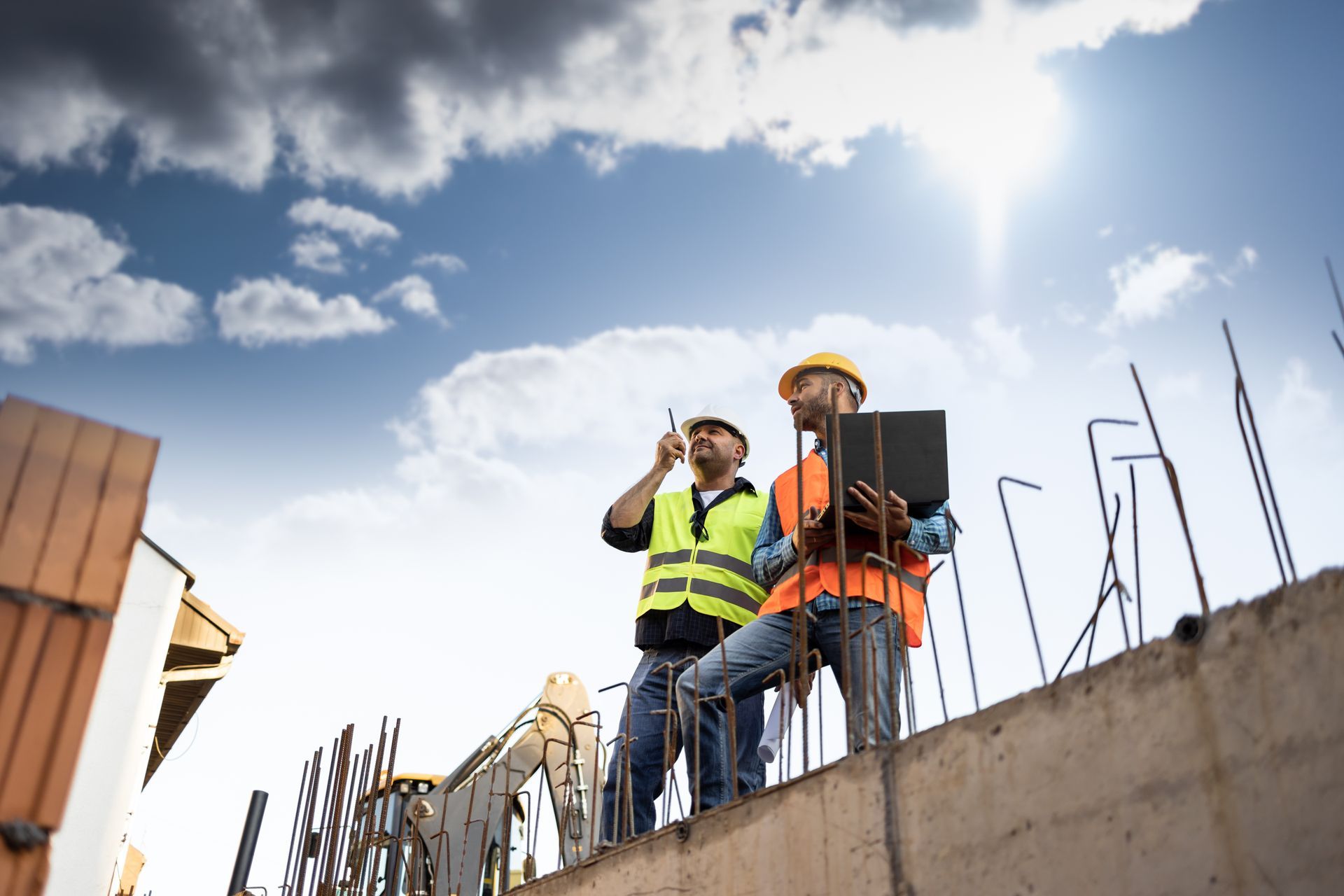Des ouvriers du bâtiment travaillent sur une structure en béton, l'un utilise une radio, l'autre tient une tablette, sur fond de ciel ensoleillé.