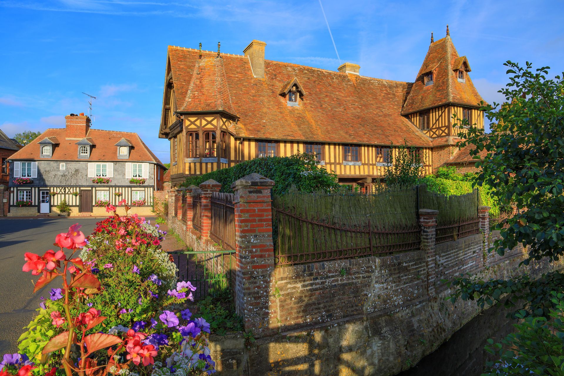 Bâtiments historiques à colombages avec toits de tuiles orange dans un village européen ; fleurs au premier plan, ciel bleu.