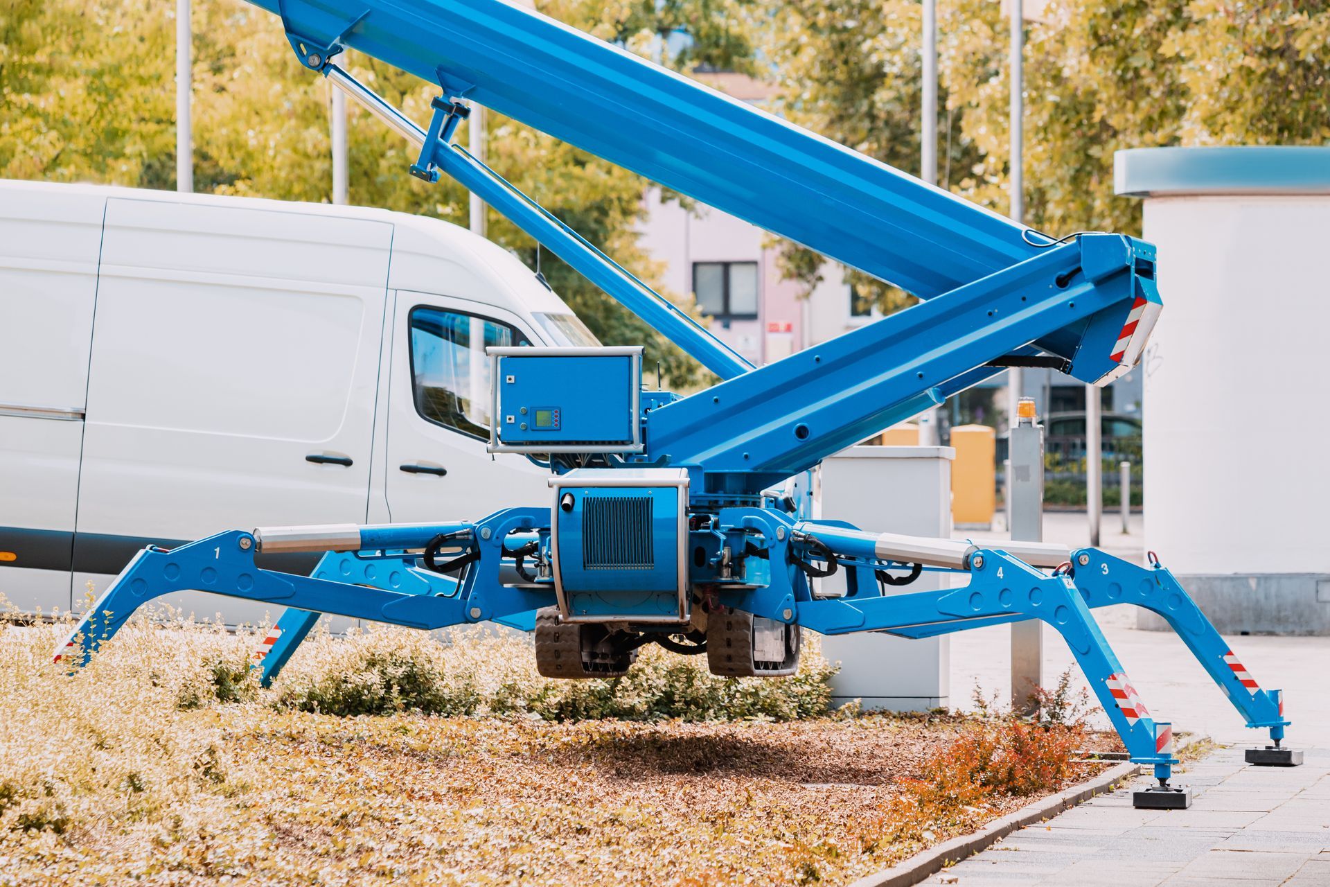 Une nacelle élévatrice bleue en forme d'araignée, aux pattes déployées, est stationnée près d'une camionnette blanche, prête à élever du personnel.