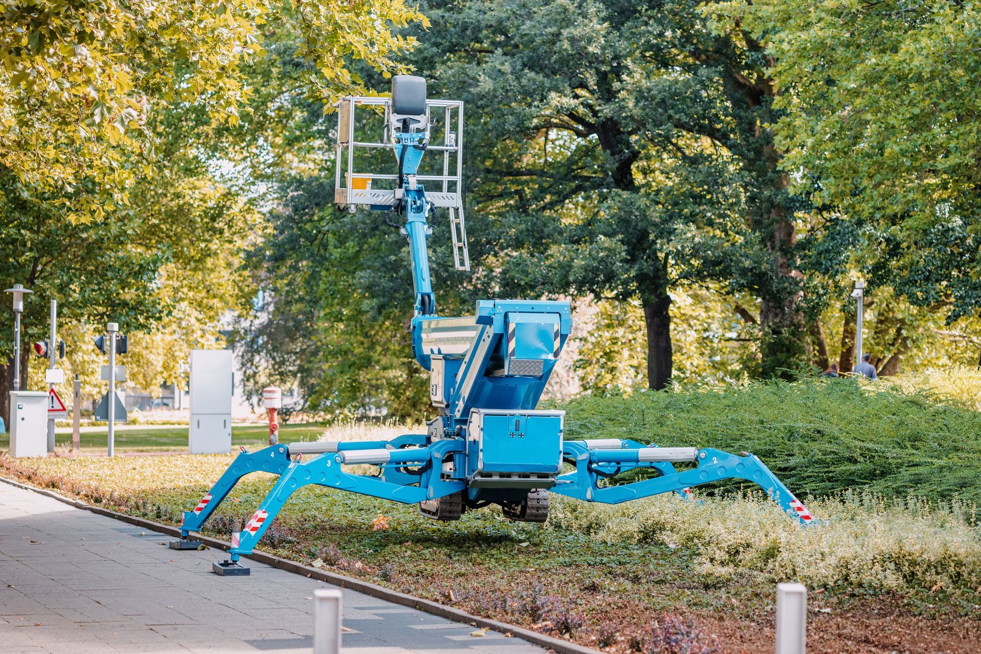 Une nacelle élévatrice articulée bleue se trouve sur un trottoir, son bras déployé vers les arbres.