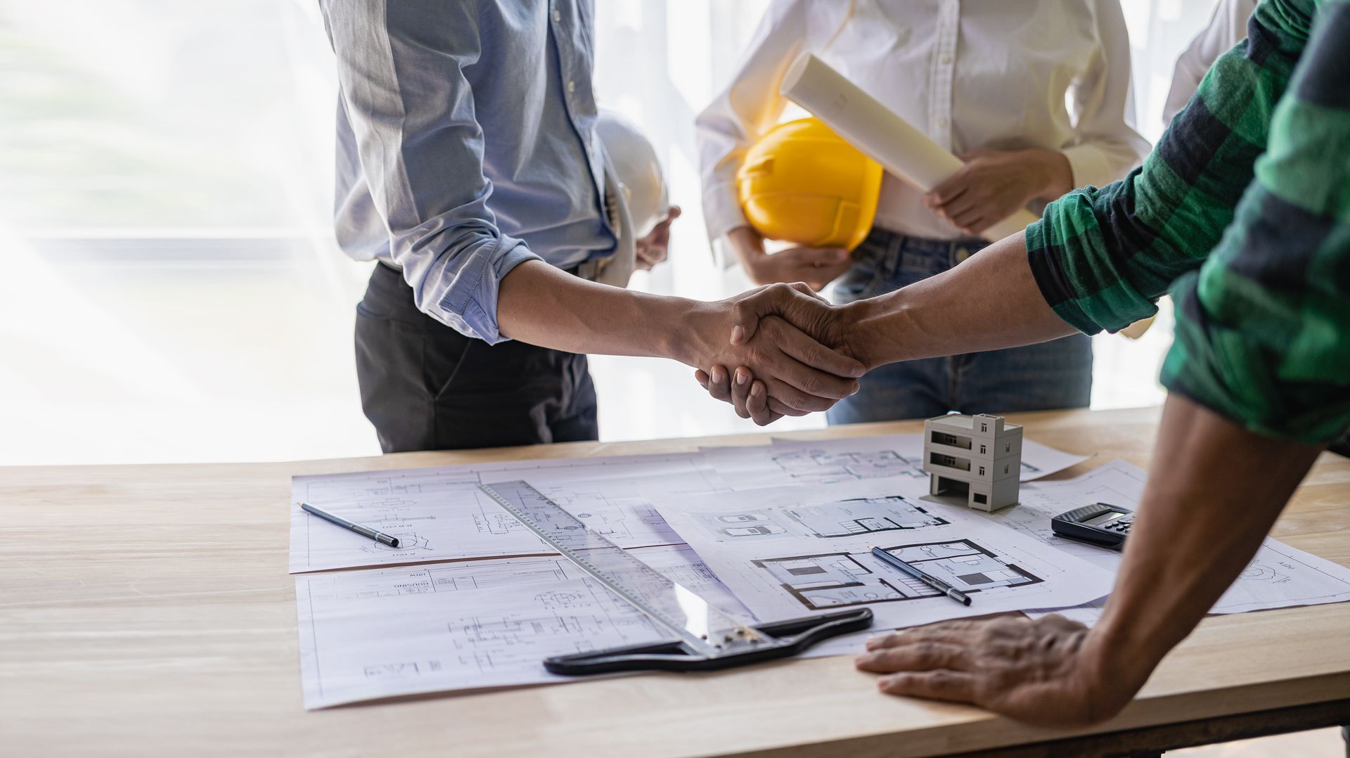 Des personnes se serrent la main au-dessus de plans disposés sur une table, tandis que des outils de construction et des casques de chantier sont visibles.