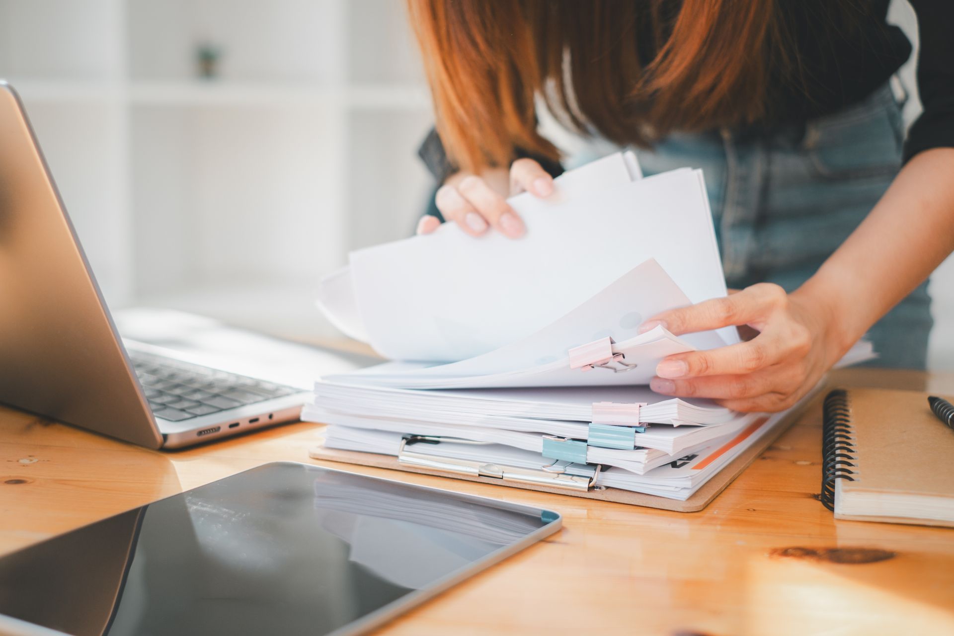 Une femme trie des documents sur un bureau, un ordinateur portable et une tablette à la main.