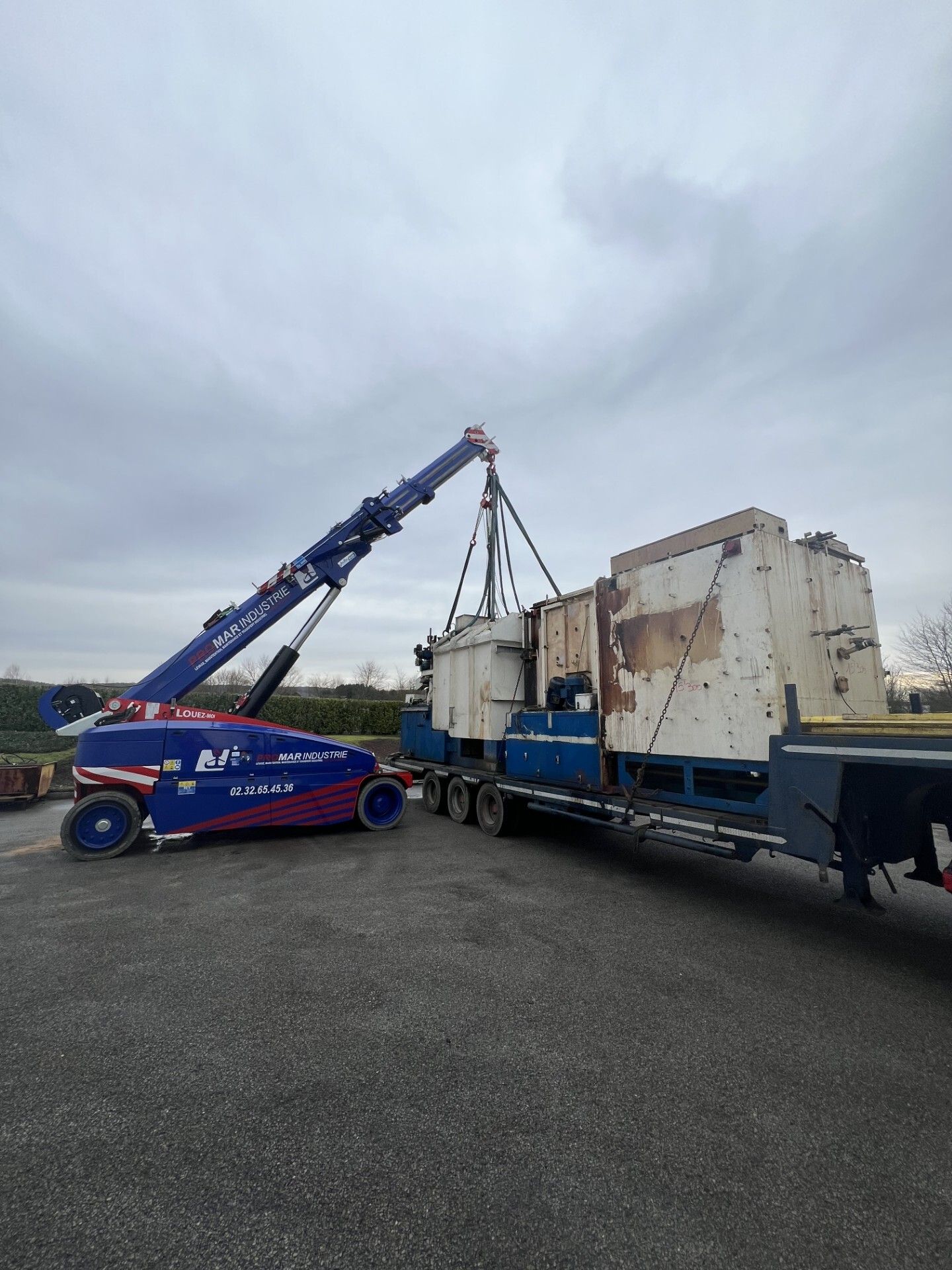 Une grue bleue soulève une grande structure en béton sur un camion plateau dans une zone industrielle.