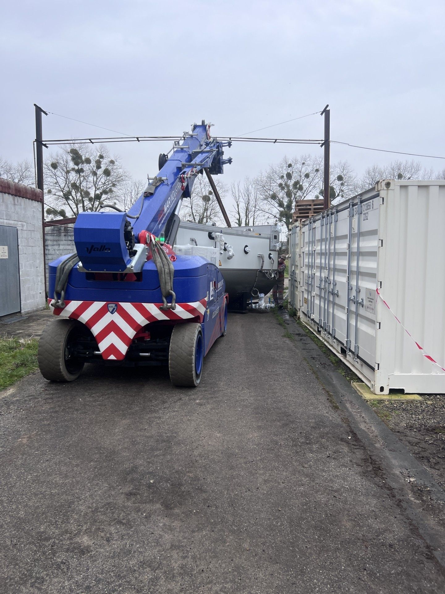Camion-grue bleu sur une voie étroite entre des conteneurs maritimes blancs.