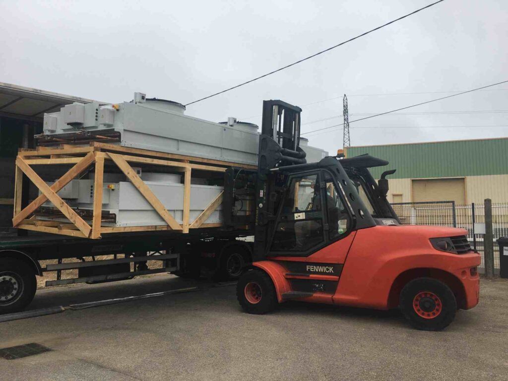 Un chariot élévateur décharge un gros composant industriel emballé dans une caisse en bois d'un camion plateau par temps couvert.