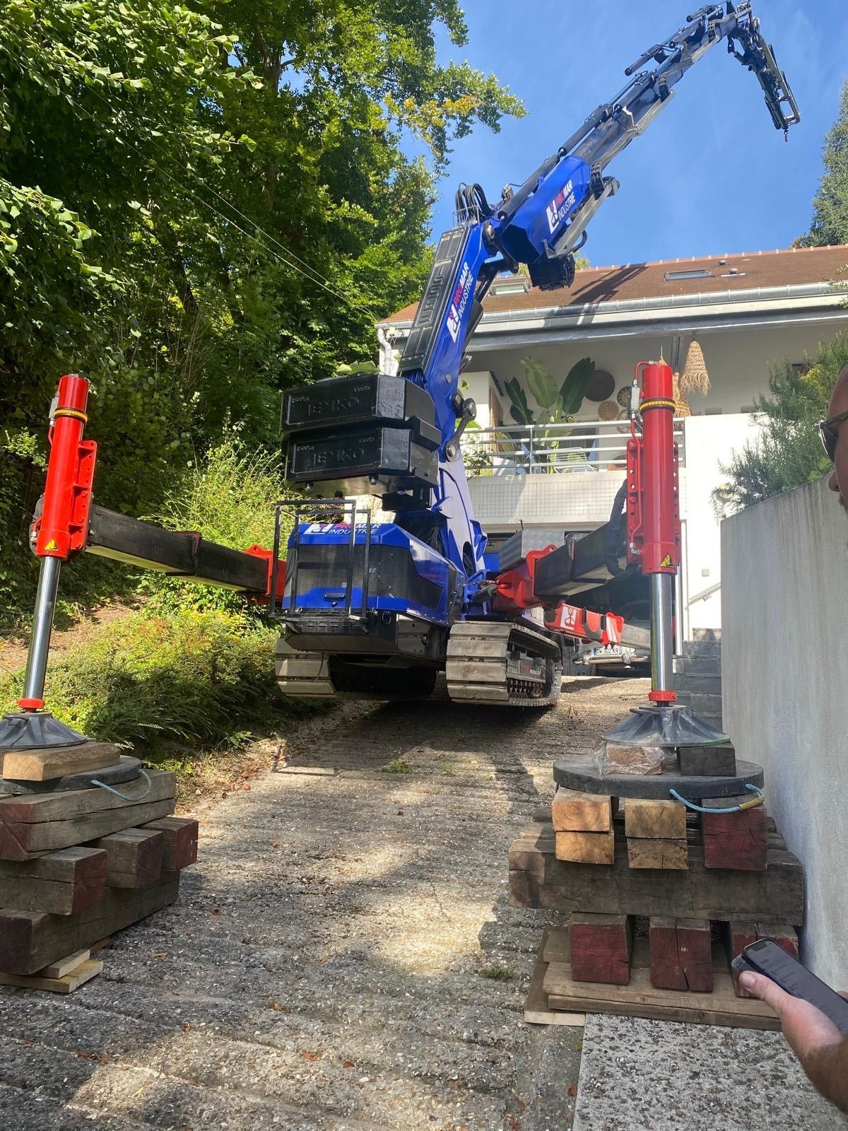 Une pelleteuse bleue soulève des débris dans une allée étroite à côté d'une maison, avec des briques empilées et des arbres à proximité.