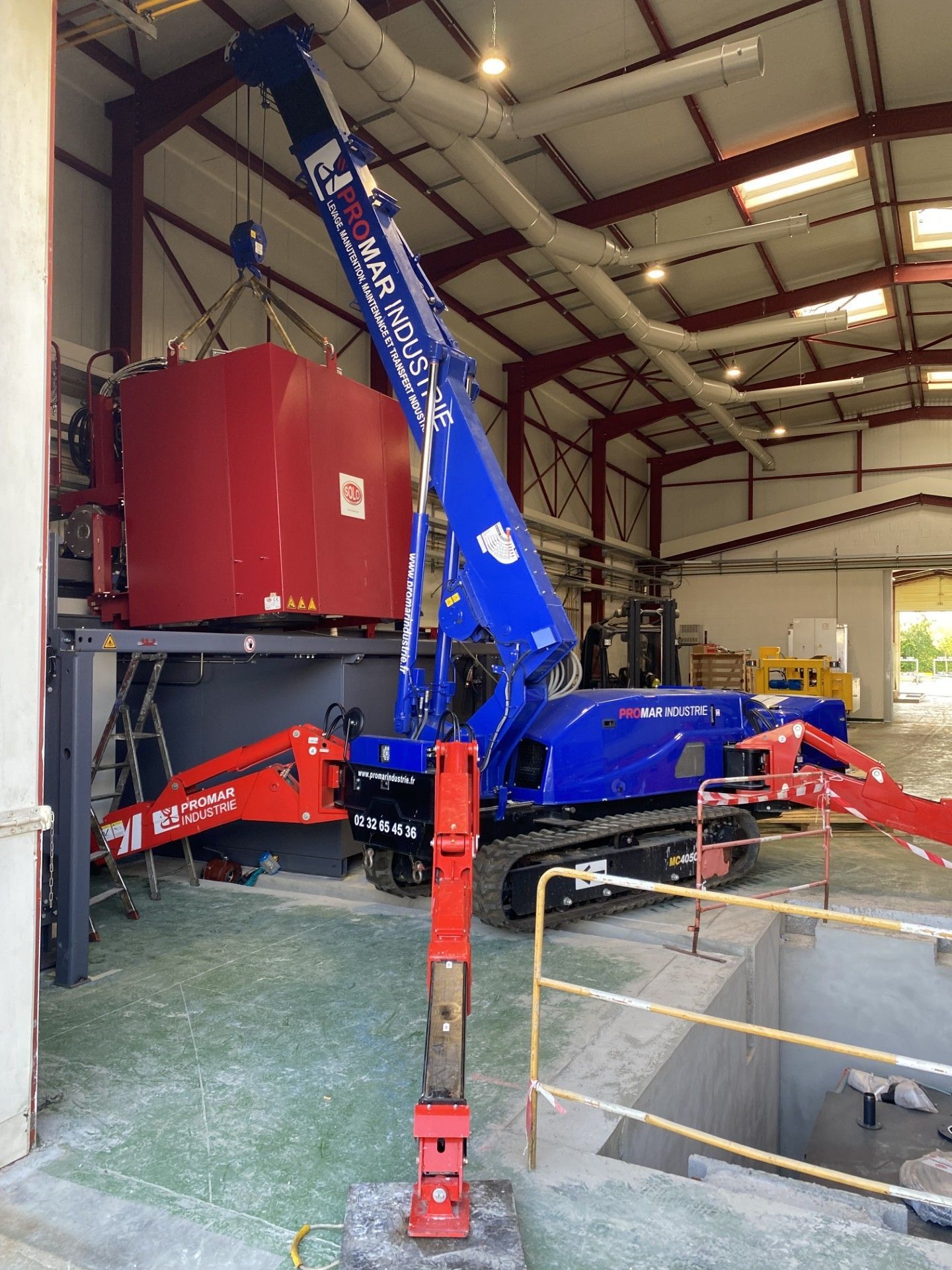 Grue mobile bleue et rouge à l'intérieur d'un entrepôt, soulevant un grand réservoir rouge près d'une porte.