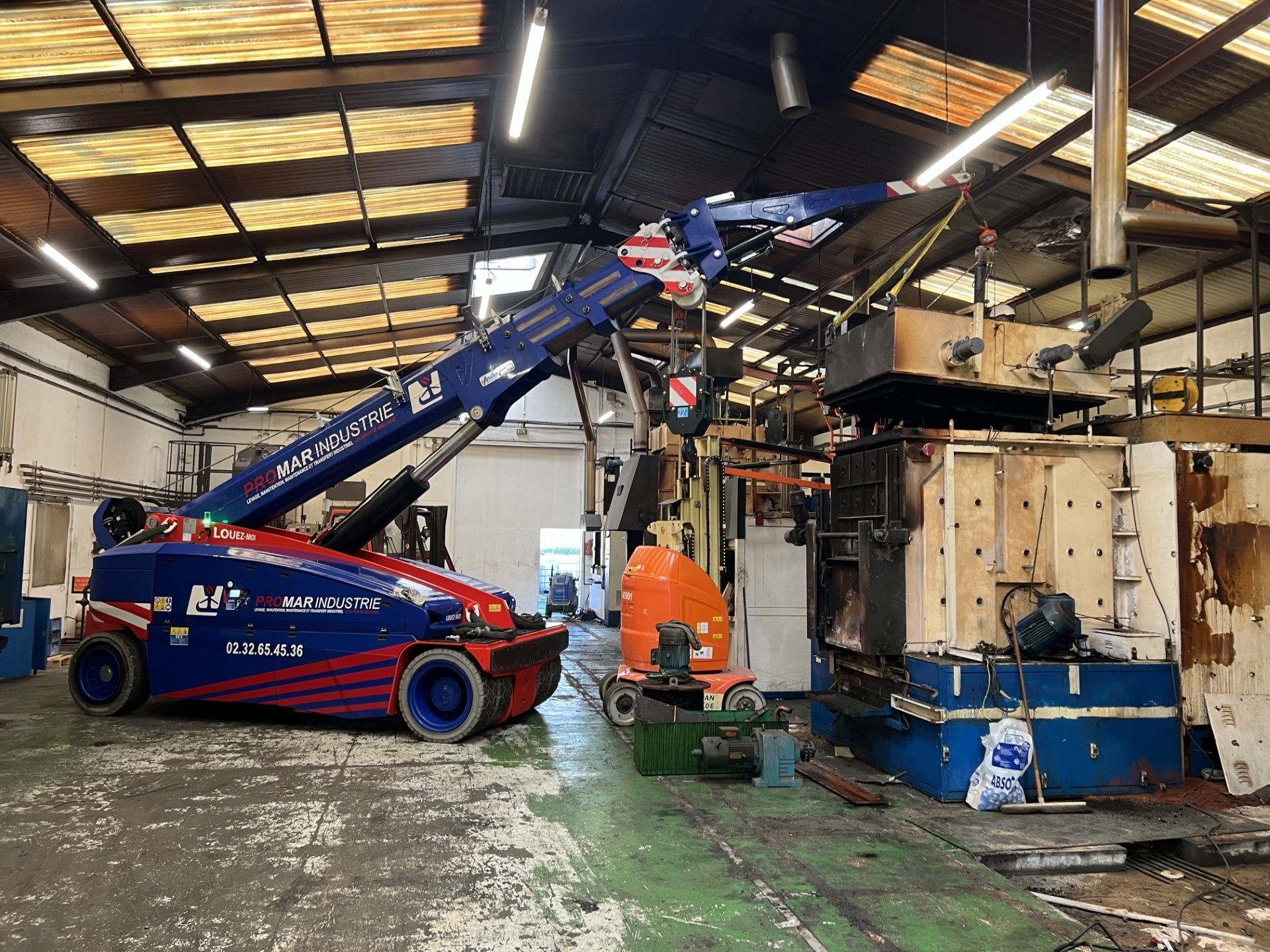 Un chariot télescopique bleu et rouge soulève une charge à l'intérieur d'un entrepôt industriel, à côté d'une grande machine.