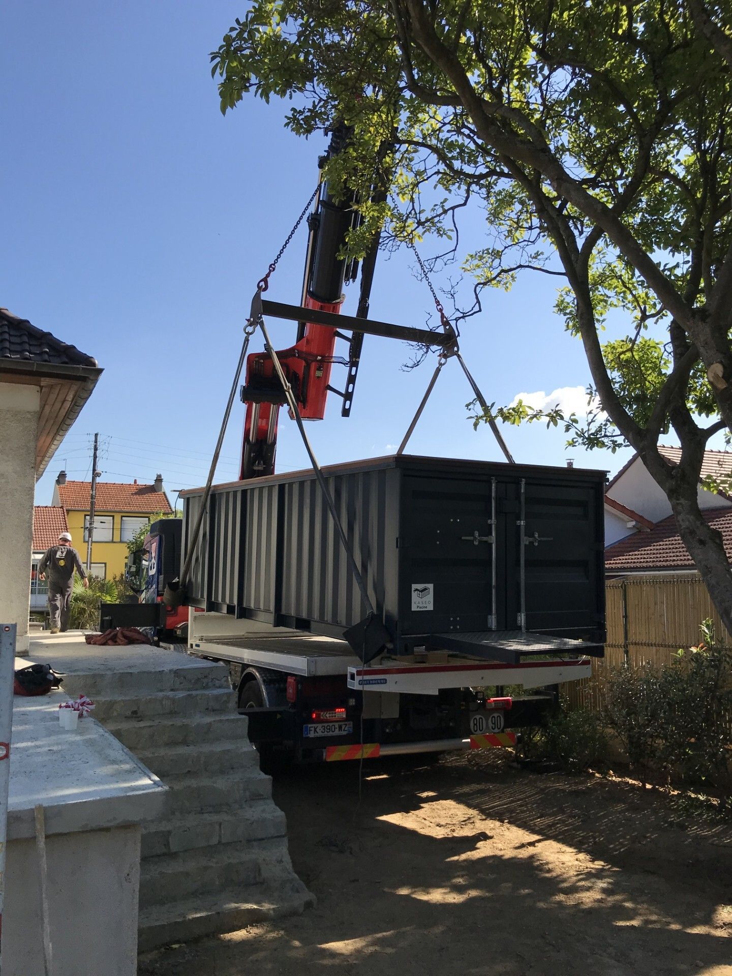 Une grue charge un grand conteneur noir sur un camion plateau dans une rue étroite.