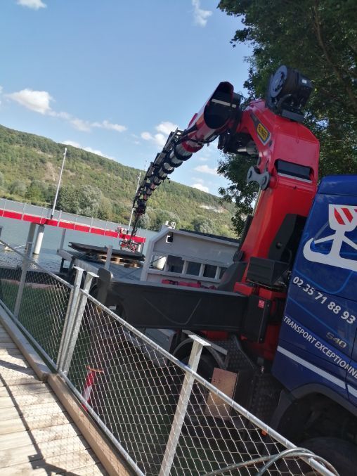 Un camion-grue rouge décharge des matériaux le long d'une allée clôturée, avec des collines verdoyantes en arrière-plan.