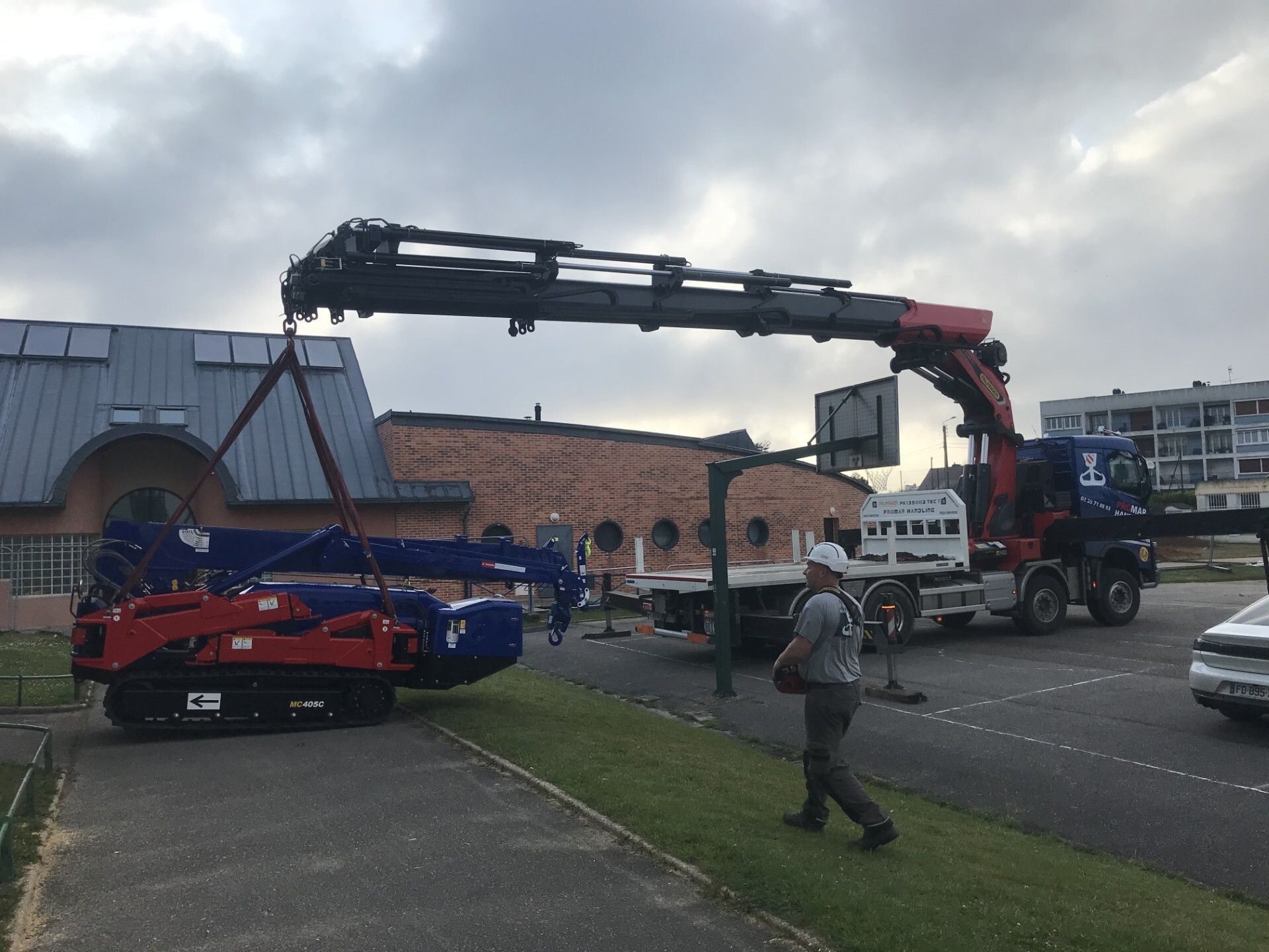 Une grande grue soulève une voiture bleue et la charge sur un camion dans un parking à côté d'un bâtiment en briques.
