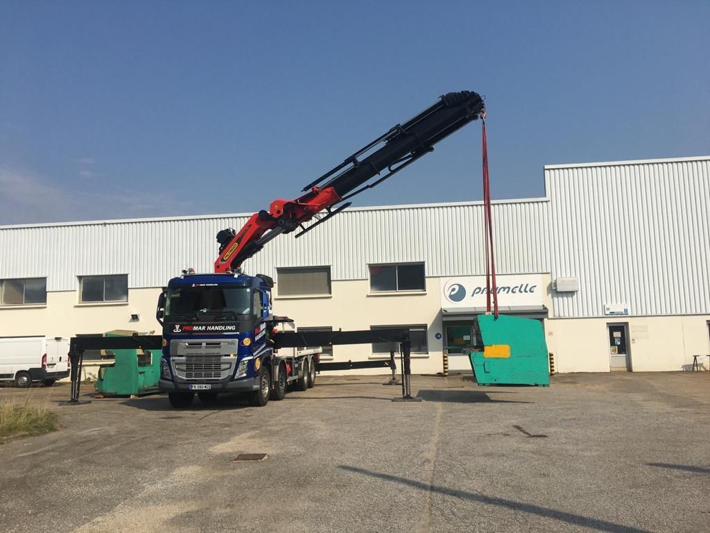 Camion-grue soulevant une poutre rouge dans une zone industrielle à côté d'un entrepôt