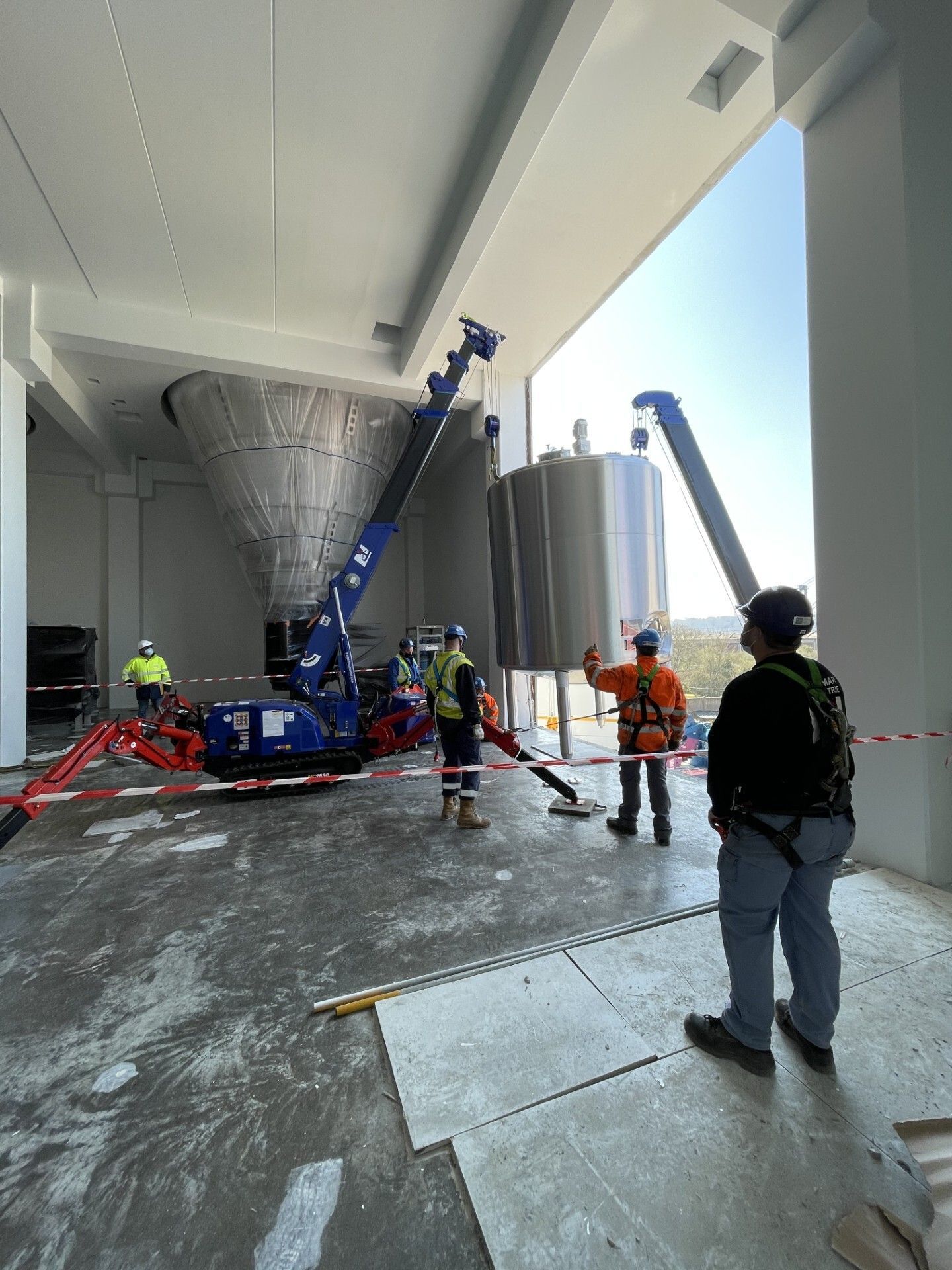 Des ouvriers inspectent une grande grue bleue à l'intérieur d'un entrepôt industriel, des cônes et des tuyaux étant posés au sol.