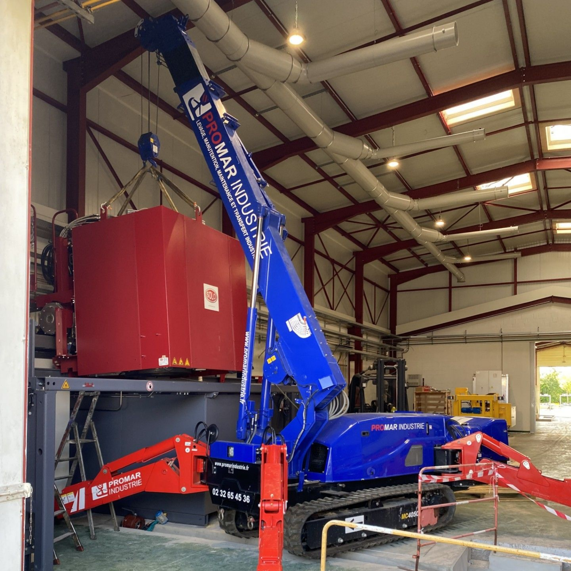 Une grue industrielle bleue et rouge soulève une grande machine rouge à l'intérieur d'un entrepôt
