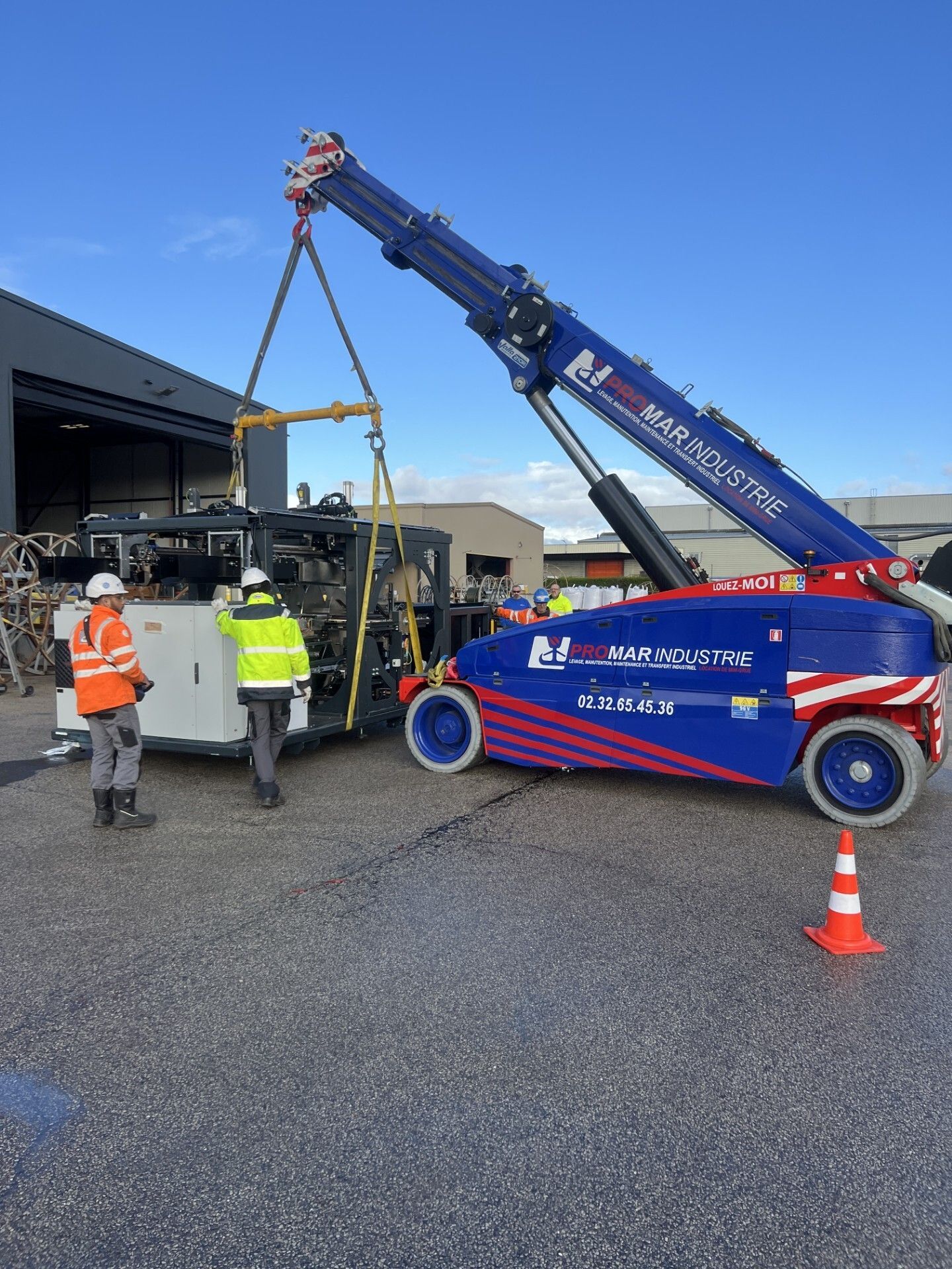 Une dépanneuse soulève un chariot élévateur sur un site industriel, avec deux ouvriers à proximité et un cône de signalisation devant.