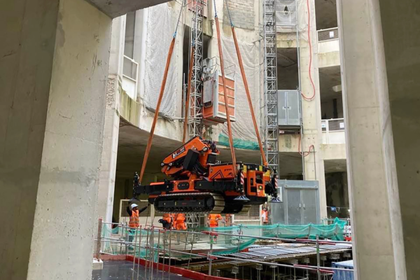 Chantier. Une grue soulève un véhicule chenillé orange à travers des ouvertures dans le béton. Des ouvriers se trouvent en dessous.