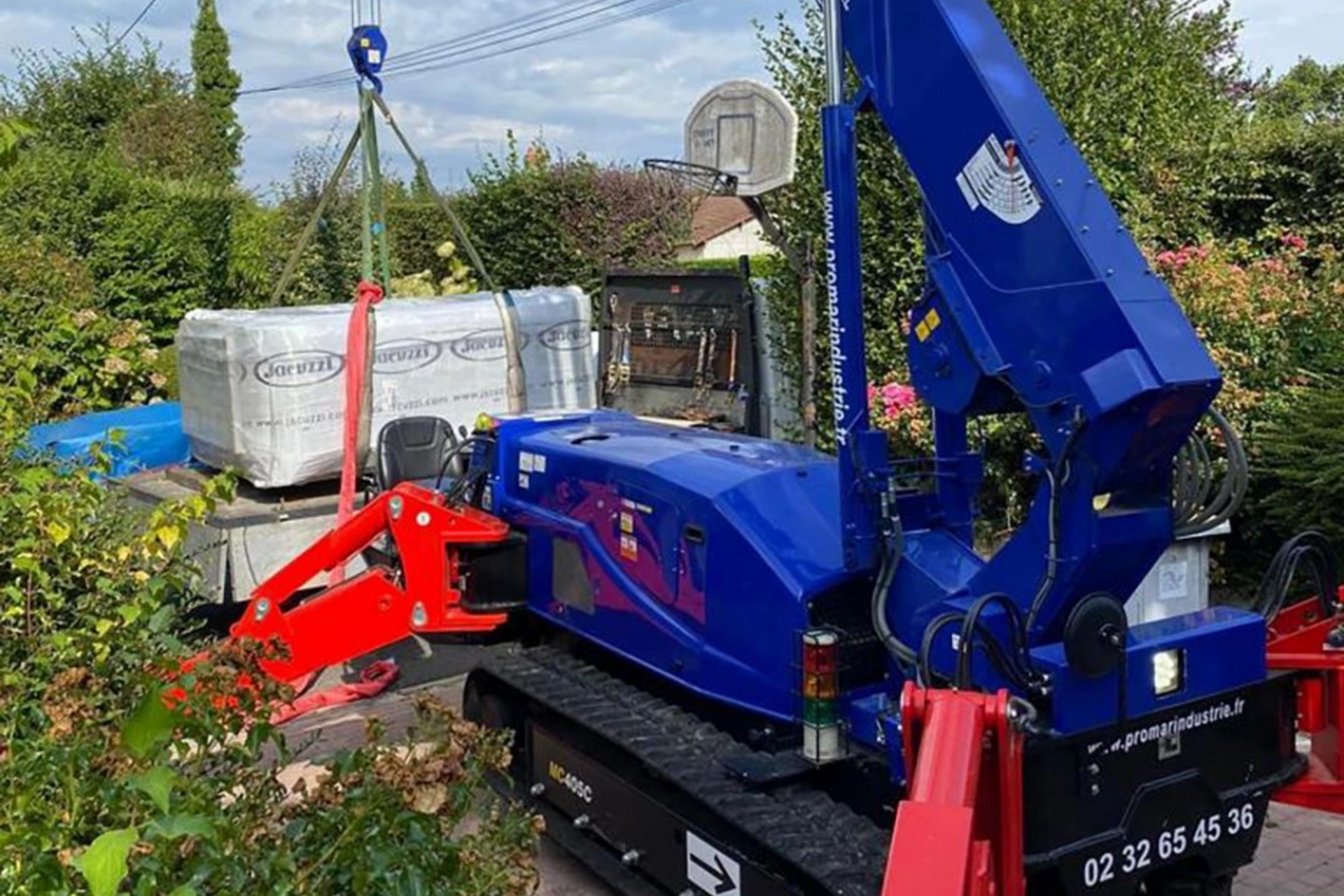 Une mini-grue bleue soulève une palette de matériaux de construction blancs d'un camion dans une cour arrière résidentielle.