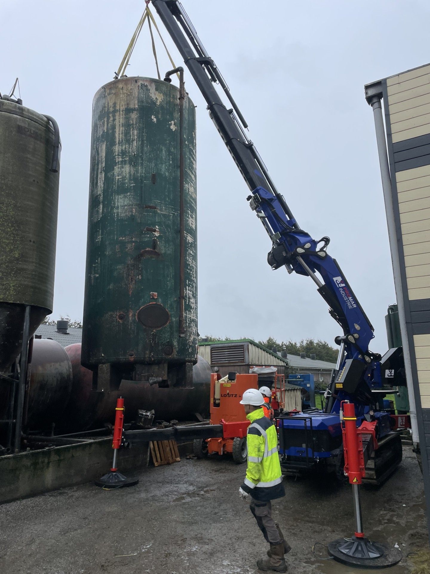 Une grue soulève un grand réservoir cylindrique sur un chantier de construction, des ouvriers portant des équipements de sécurité se trouvant à proximité.