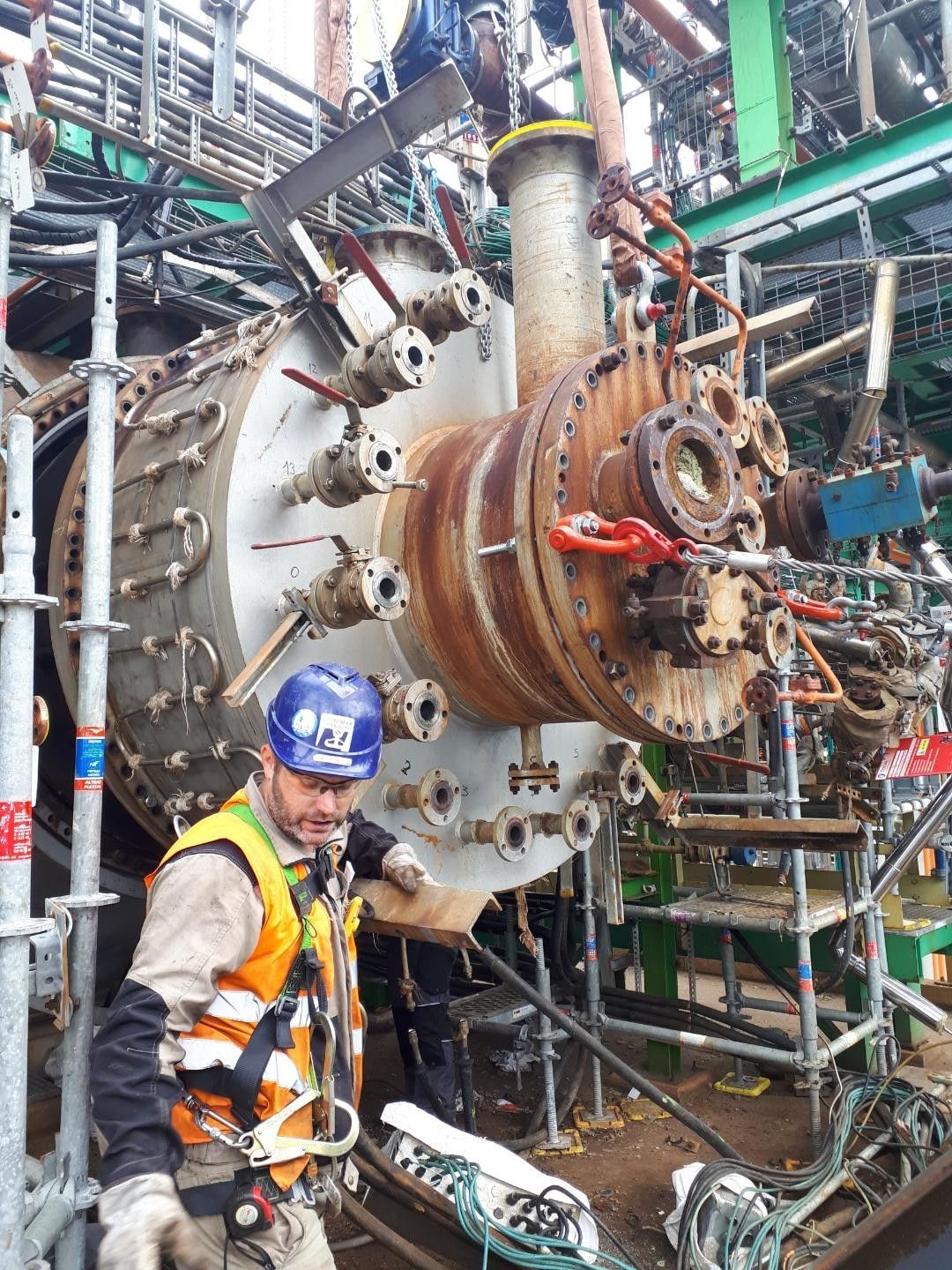 Ouvrier portant un casque de chantier à côté d'une grande machine industrielle dans une usine en activité, avec des échafaudages et des canalisations au-dessus de sa tête.