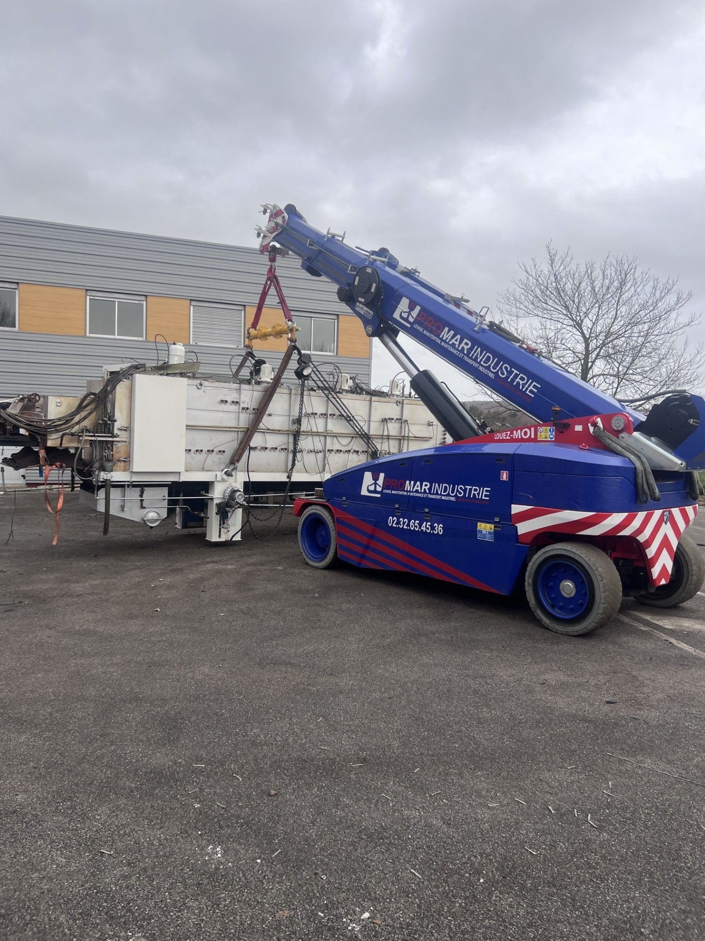Un camion-grue bleu soulève une structure blanche dans une zone industrielle sous un ciel nuageux.