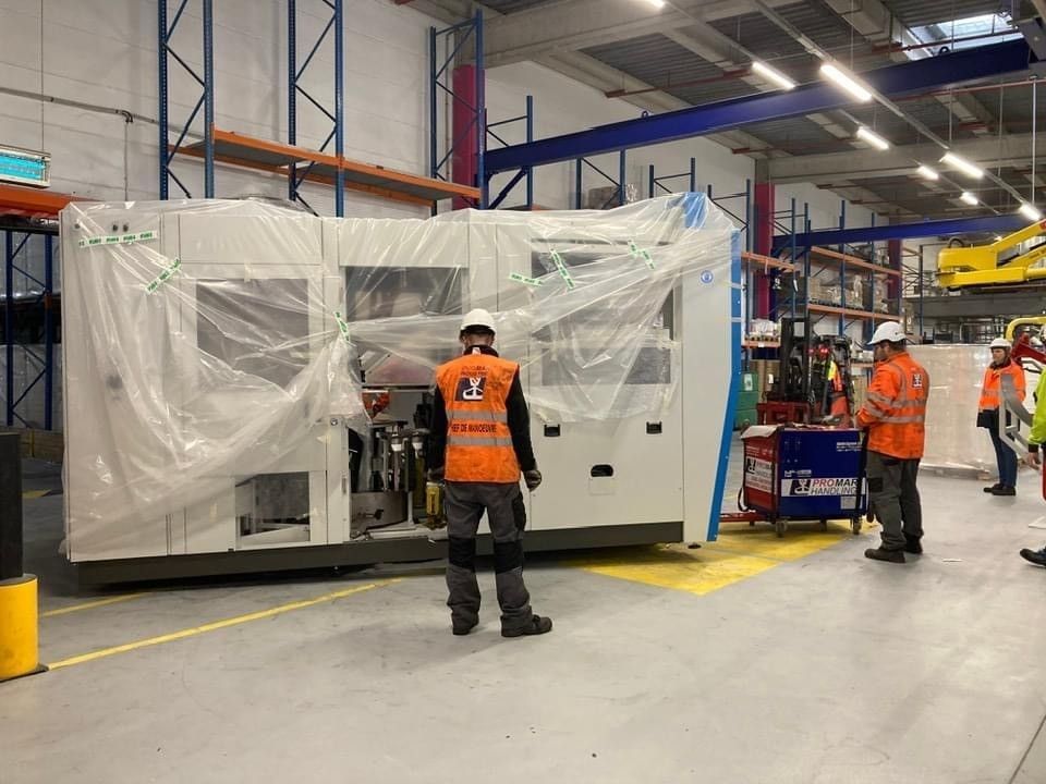 Des ouvriers vêtus de gilets orange inspectent une grosse machine emballée dans du plastique à l'intérieur d'un entrepôt.
