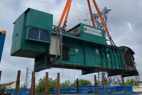 Grosse machine industrielle verte suspendue par des sangles orange ; à l'extérieur, près de lignes électriques.