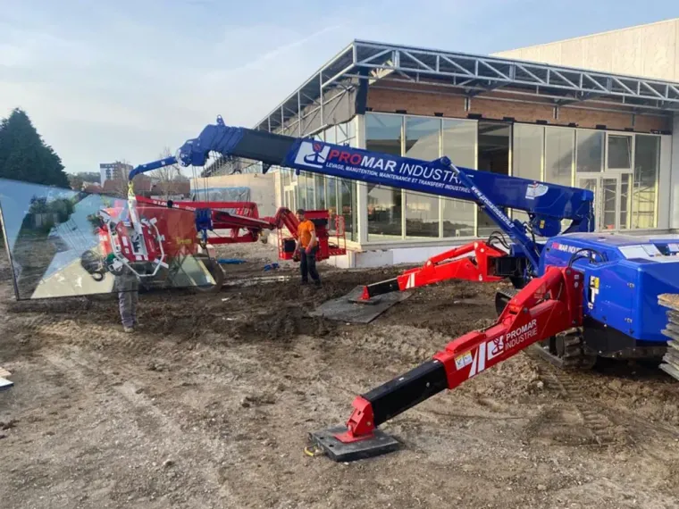 Deux mini-grues soulèvent des panneaux de verre à l'extérieur d'un bâtiment ; sol boueux. Rouge et bleu.