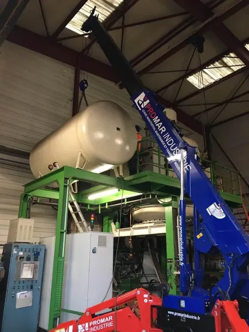 Une grue soulève un grand réservoir cylindrique sur une plateforme métallique verte à l'intérieur d'un bâtiment.