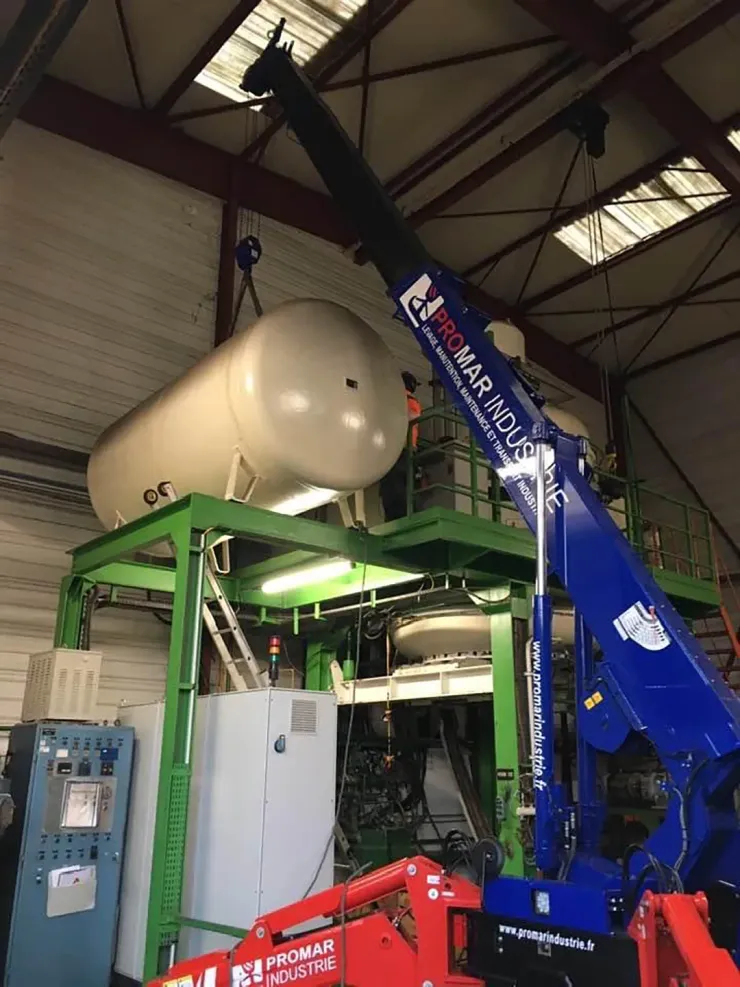 Une grue soulève un grand réservoir cylindrique et le dépose sur une structure métallique verte à l'intérieur d'un bâtiment.