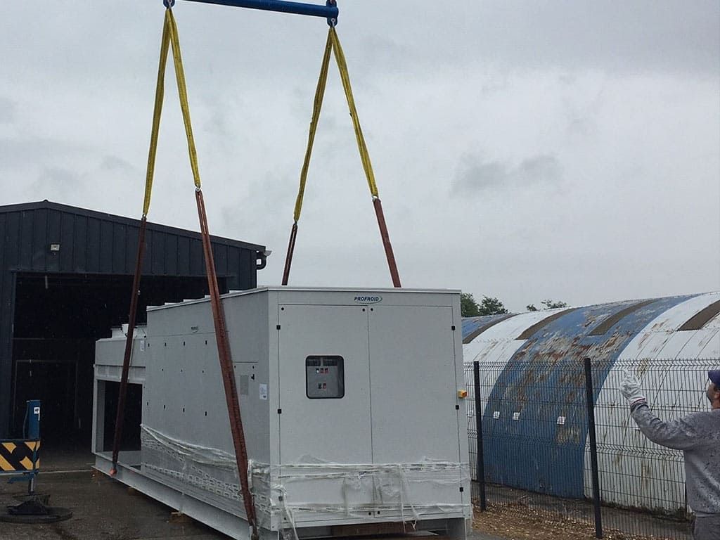 Un gros générateur est soulevé par une grue à l'aide de sangles jaunes devant un bâtiment.