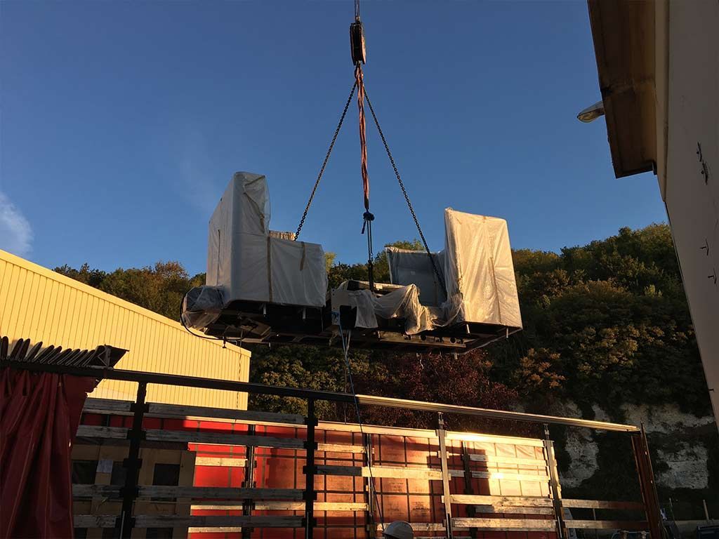 Une grue soulève deux gros composants industriels recouverts d'une bâche, probablement en vue de leur installation. En extérieur, de jour.