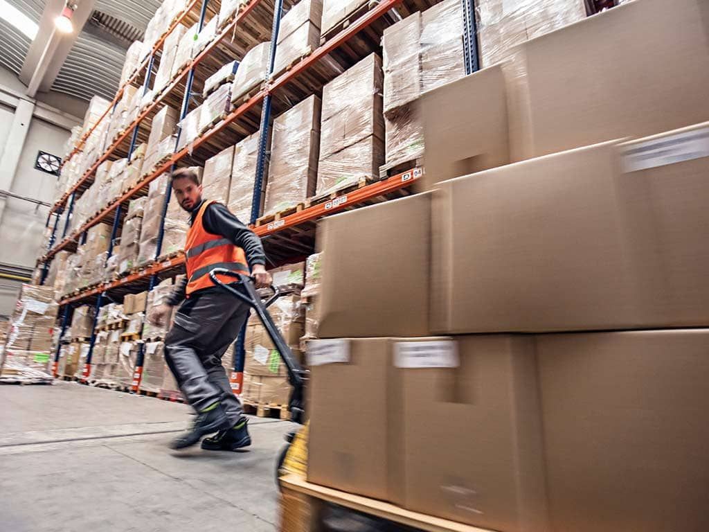 Ouvrier d'entrepôt portant un gilet de sécurité tirant un transpalette chargé de cartons.