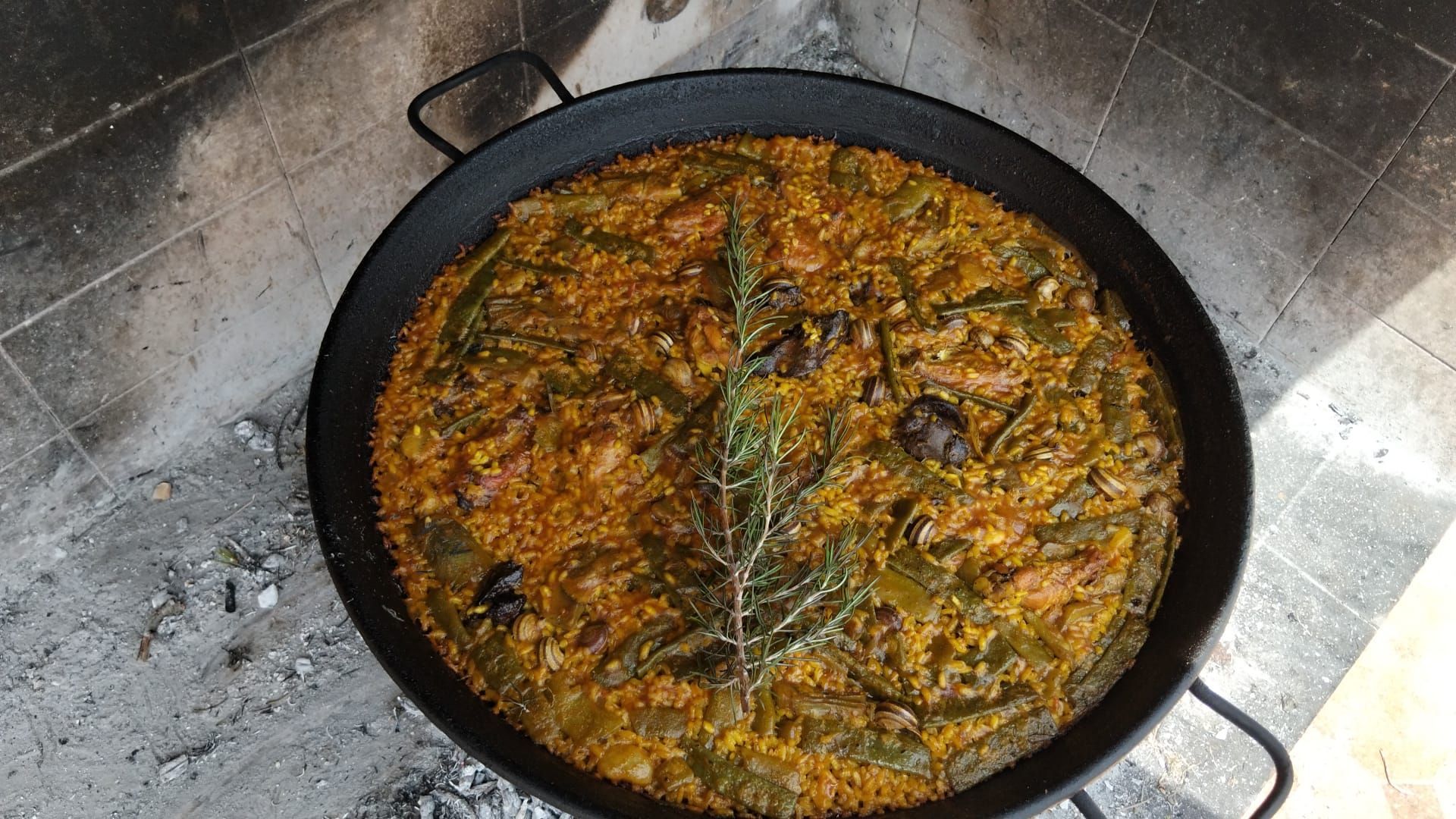 Paella en una gran sartén negra cocinándose sobre un pozo de fuego, con judías verdes y hierbas.