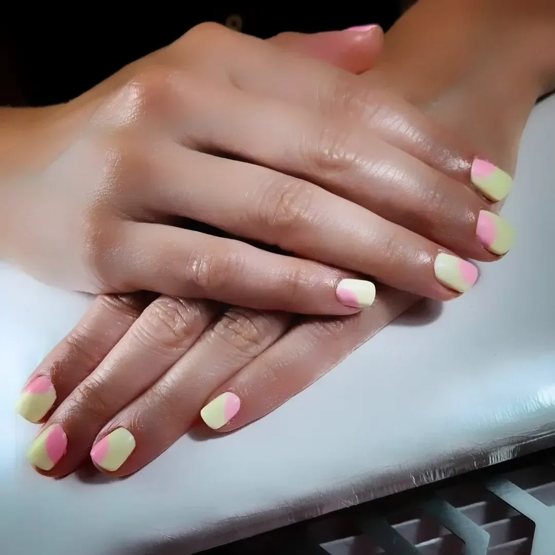 Primer plano de manos con uñas pintadas en tonos amarillo pastel y rosa, apoyadas sobre una superficie blanca.